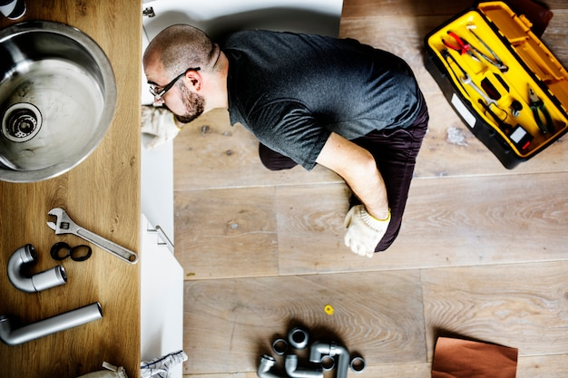 A plumber kneeling under a sink, surrounded by tools and pipes, while fixing plumbing issues.