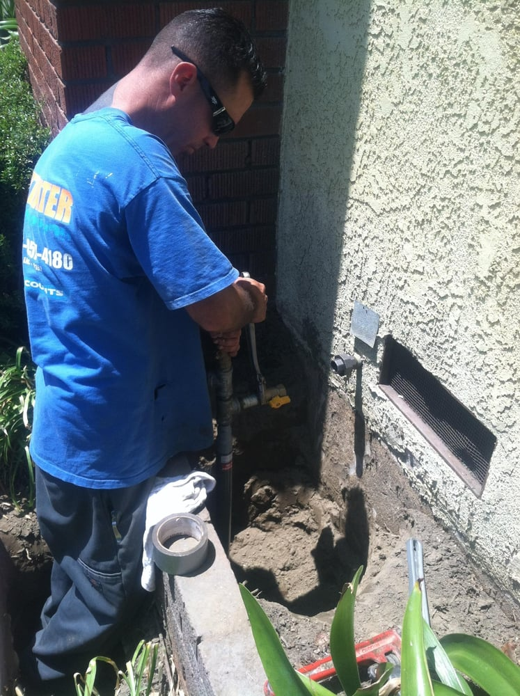 Plumber working on pipes beneath a sink with tools and water lines exposed.