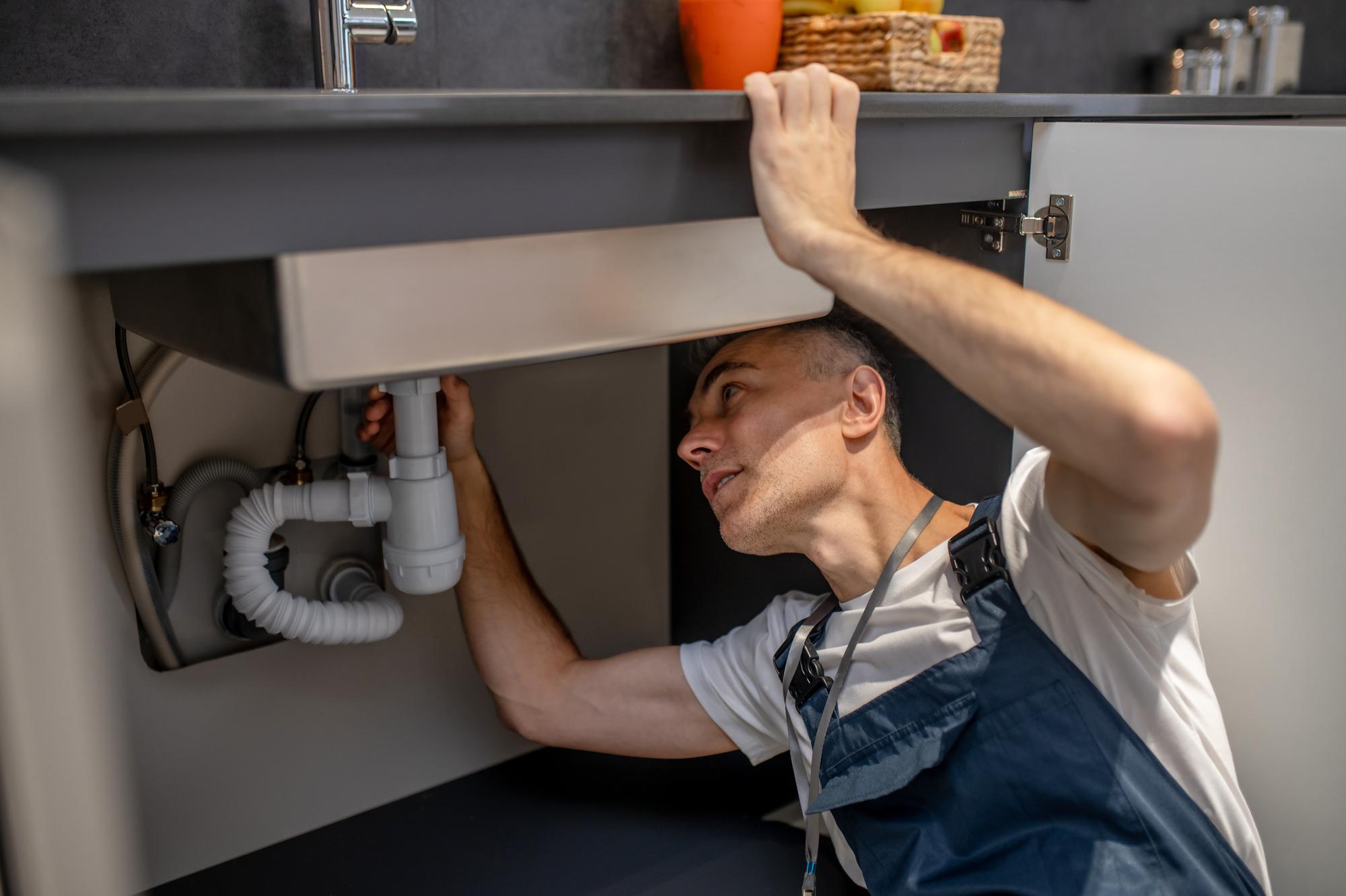 Middle-aged plumber inspecting the drain system under a kitchen sink for potential clogging issues.