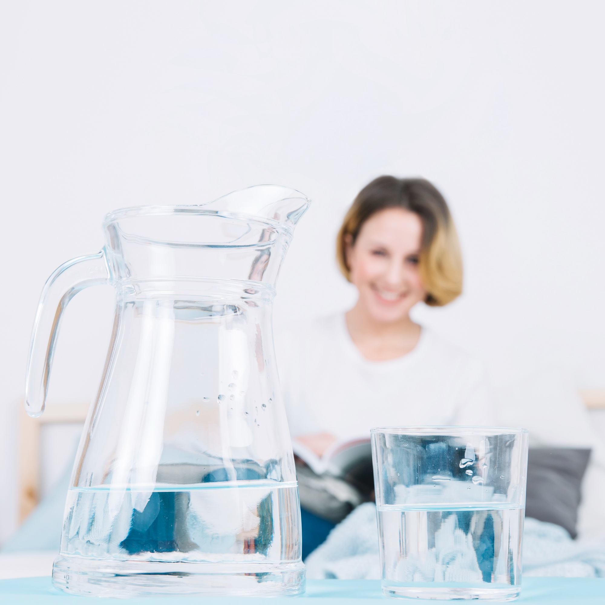 A jug and glass of clean water next to a reading woman.