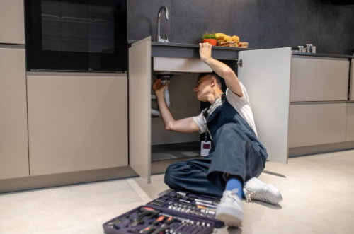 Middle-aged plumber inspecting pipe under modern kitchen sink