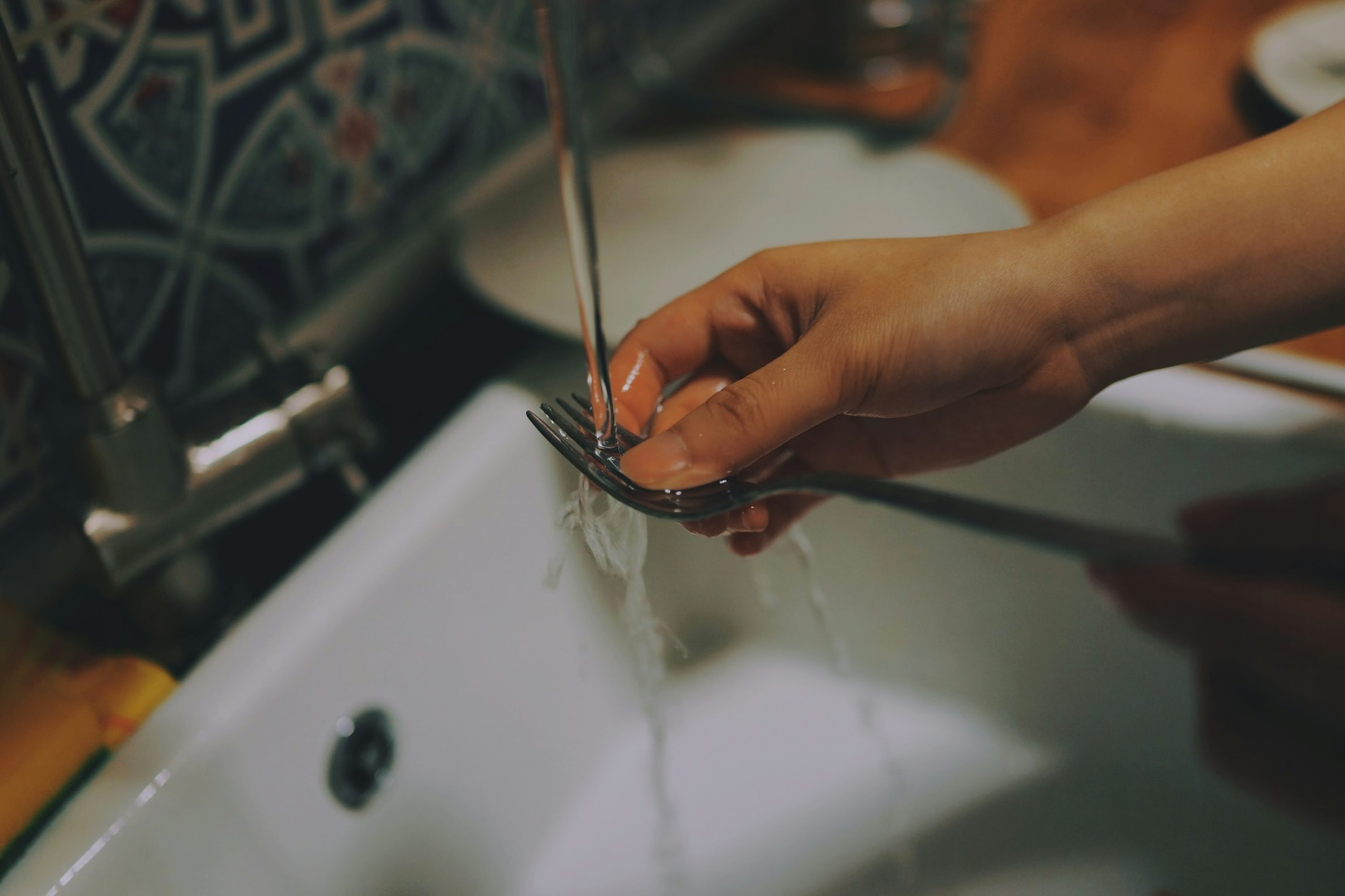 A person washing a metal fork under running water at a kitchen sink.